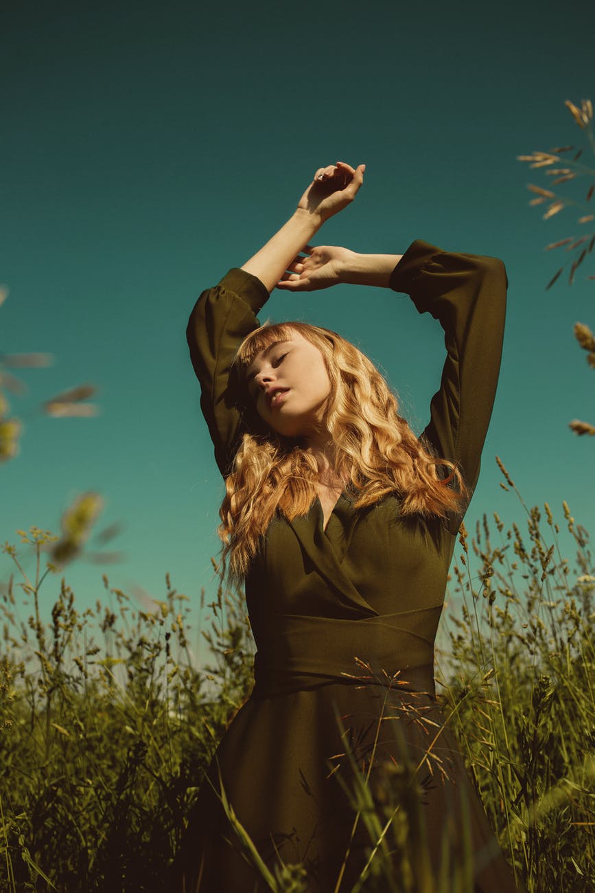 blond woman in dress standing in green meadow with arms raised in ecstasy